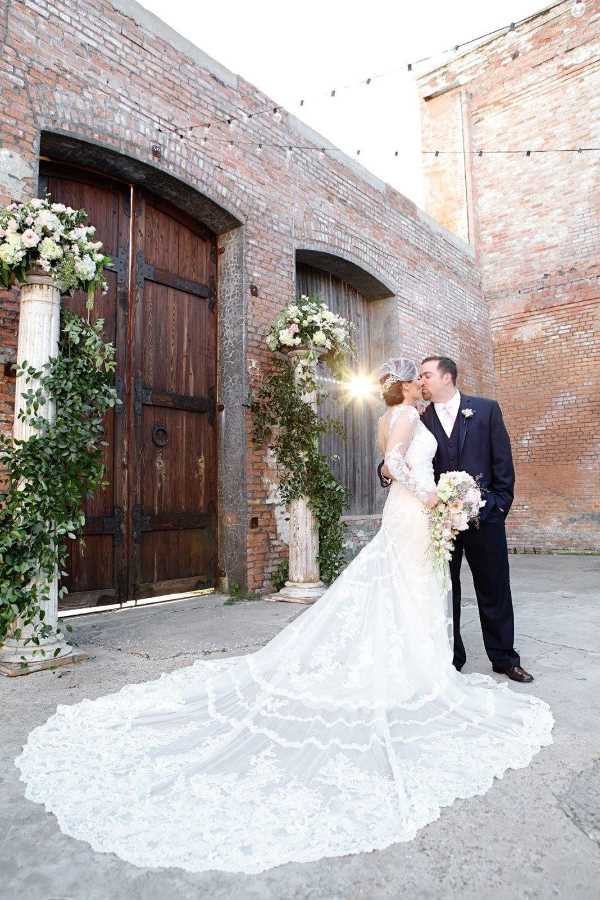 Couple kissing in front of large wooden doors and exposed brick in outdoor area.