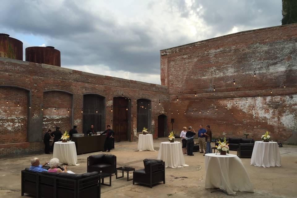 Outdoor wedding reception in open-air Dye Room at The Cotton Mill. A couple sits on a couch and there are 5 fives with two other groups of people standing.