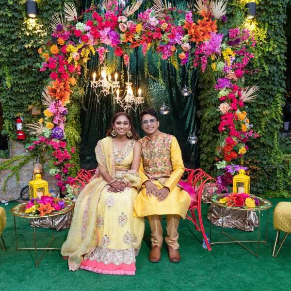 Bride and groom sitting under arch of colorful flowers.