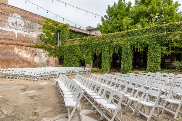 Outdoor wedding ceremony setup with white chairs and natural greenery. The brick wall has The Cotton Mill logo.