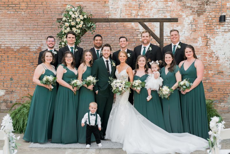Full wedding party poses in front of handcrafted wooden altar and brick wall background. 