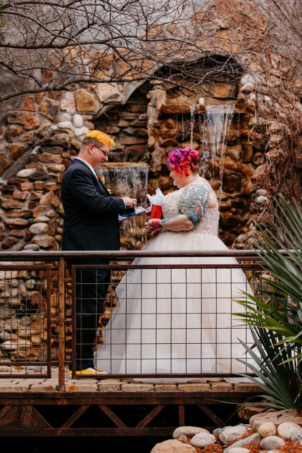 Groom giving ring to bride on bridge in front of waterfall in the outdoor courtyard area.