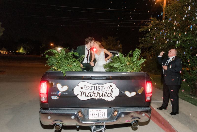 Couple kissing in bed of truck while guests shower them in bubbles. The tailgate of the truck says "Just married."