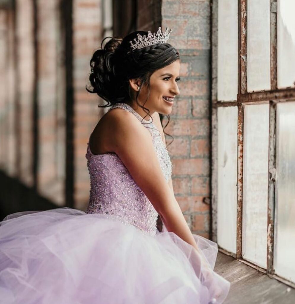 Girl in Quinceañera dress smiles and looks out industrial window.