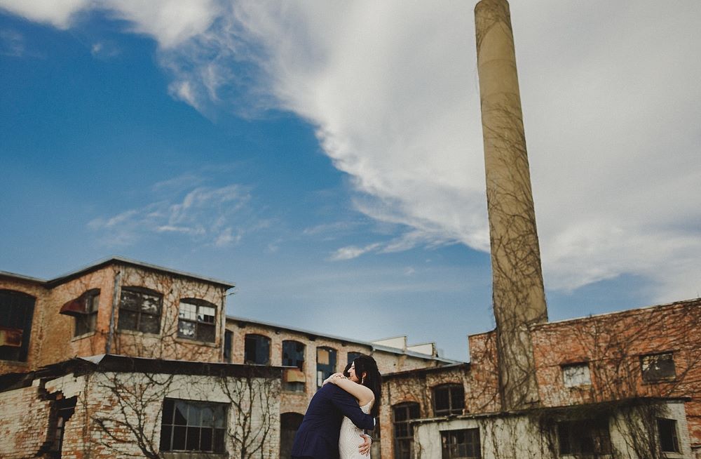 Couple hugging with The Cotton Mill smoke stack and building covered in vines behind them.