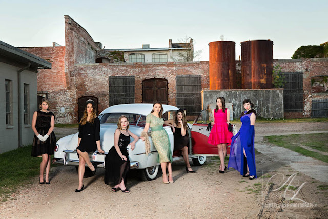 Group of women in vintage dresses pose with old car outside of The Mill.