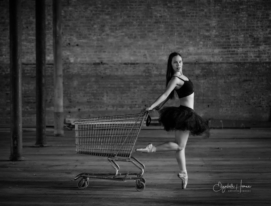 Ballerina strikes a pose with shopping cart in front of brick and wooden beams in empty warehouse space.
