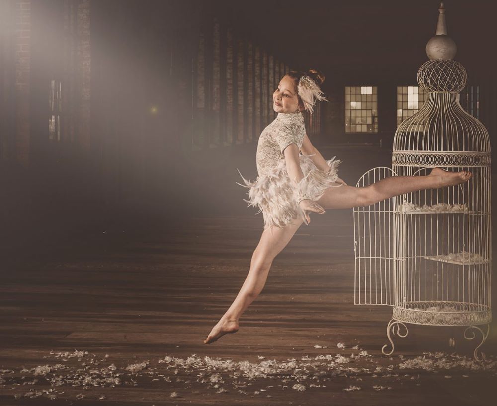 Young ballerina mid-air in front of vintage bird cage with feathers on the hardwood floor and vintage windows in the background.