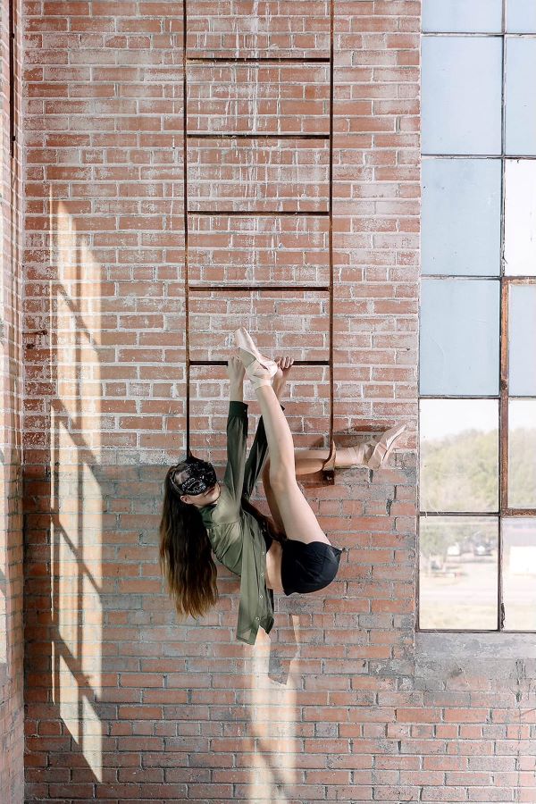 Ballerina hangs off of raised ladder in front of exposed brick. She's wearing a mask.