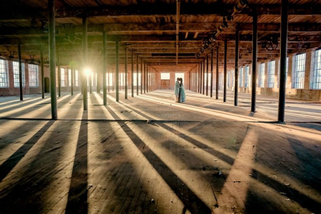 Couple inside the original weaving room as sun rays create dramatic light flares. They are far from the camera.