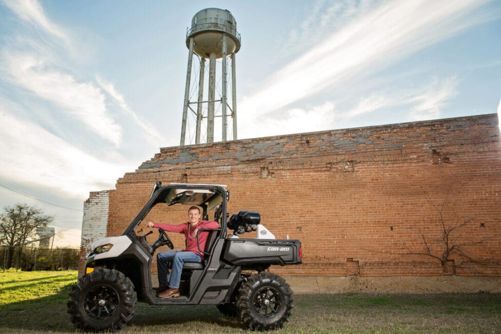 Guy in Can-Am vehicle in front of old brick and water tower.